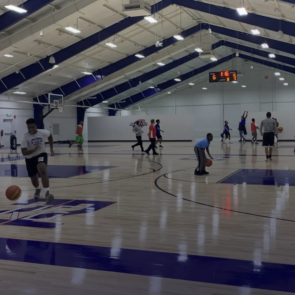 Indoor basketball court with players in various positions practicing and playing a game under a scoreboard showing 1:16 remaining in the third period.
