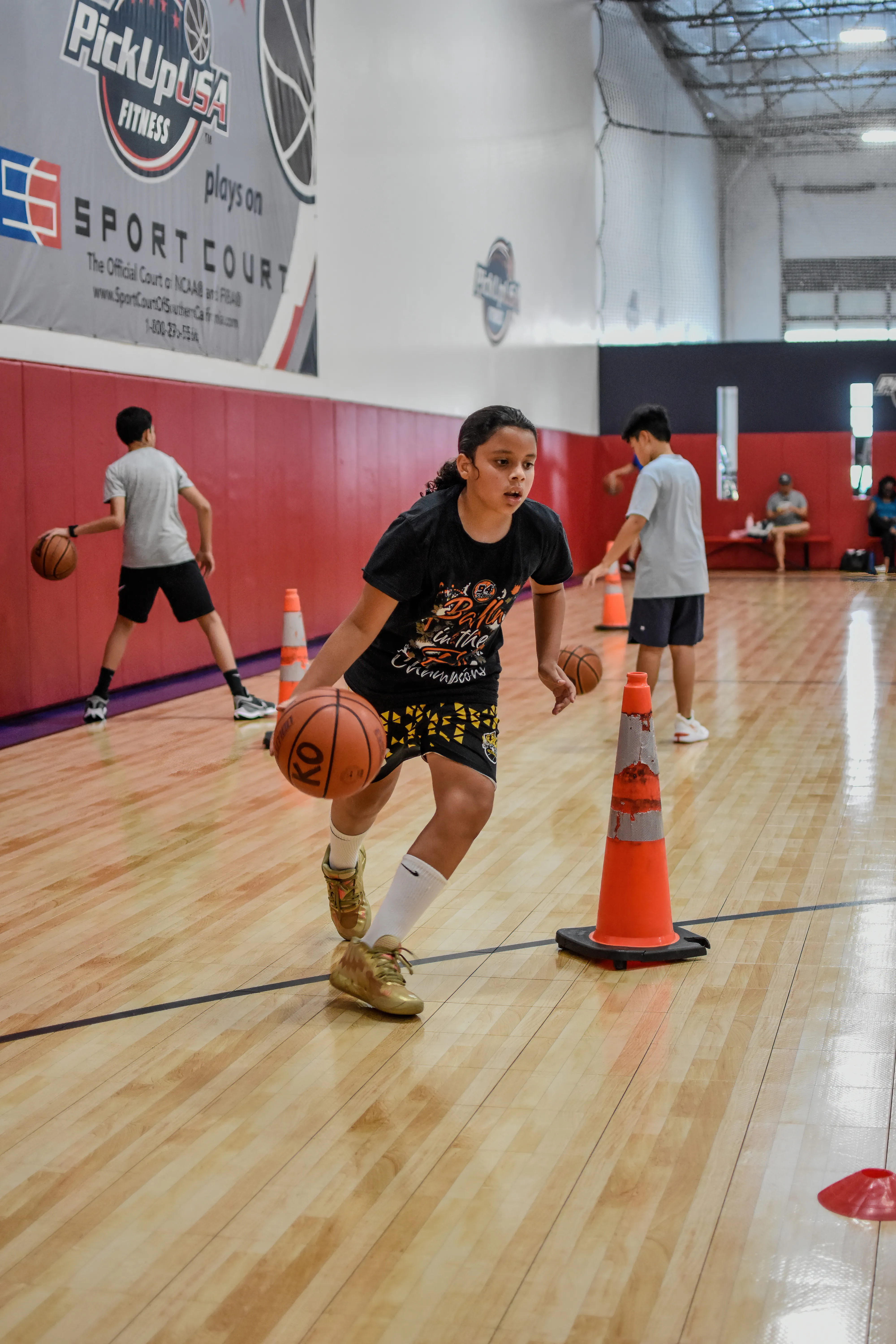 Girl dribbling a basketball around an orange cone during an indoor basketball practice with other kids in the background.