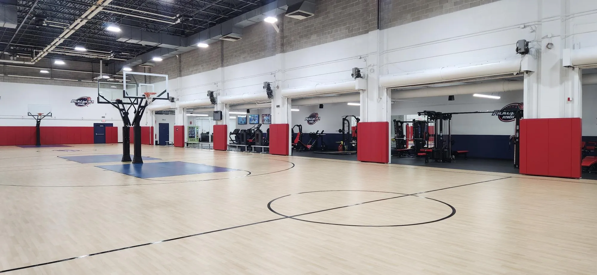 Indoor basketball court with two hoops and adjacent fitness area equipped with machines and red padding on walls.