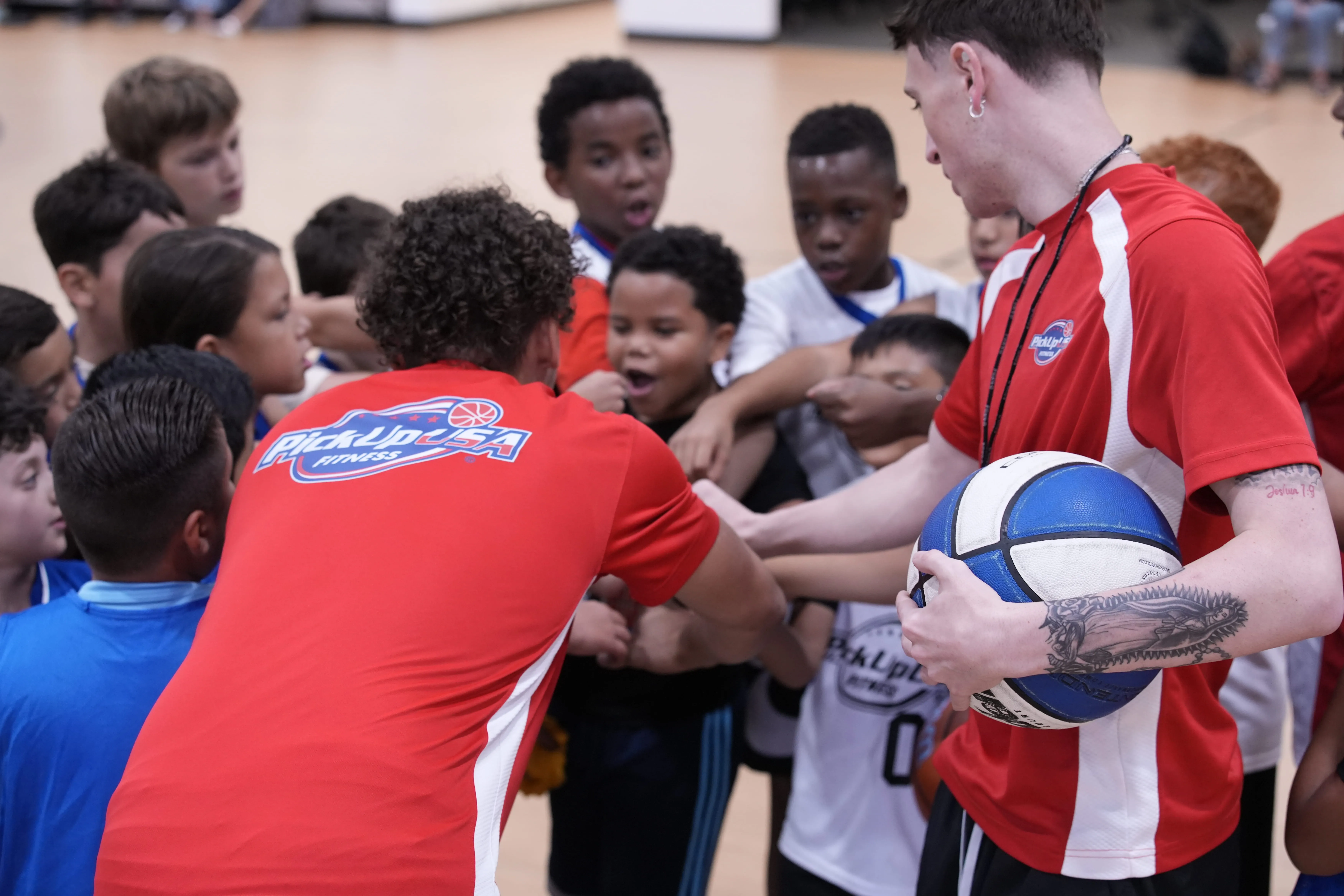 Two coaches in red PickUp USA shirts leading a group of children in a team hand huddle on an indoor basketball court.