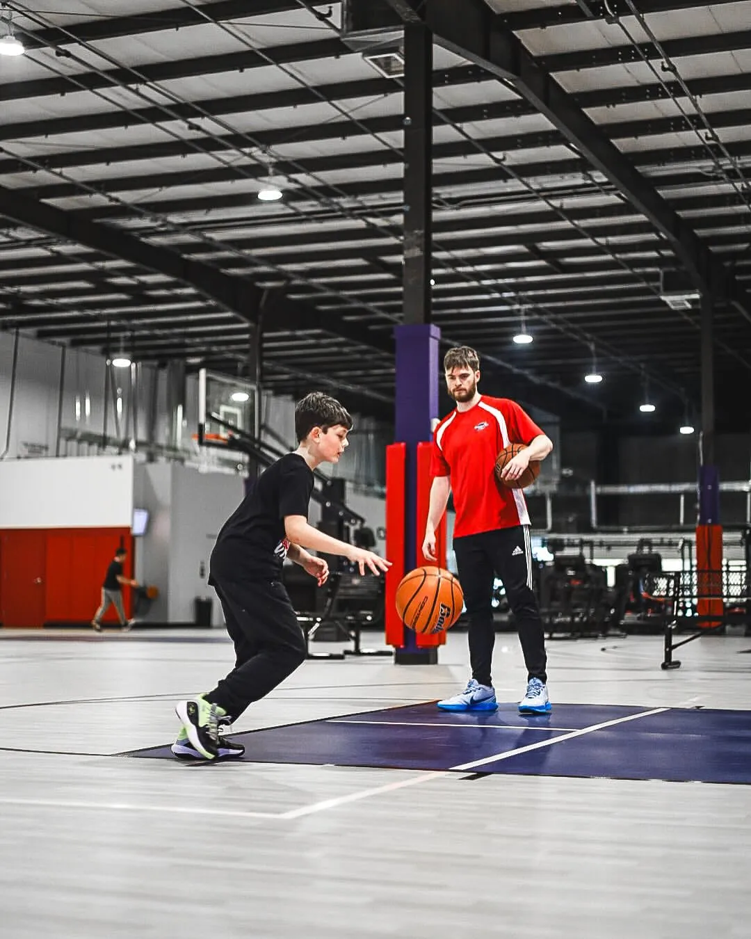 Boy dribbling a basketball during a training session with a coach holding a basketball on an indoor court.