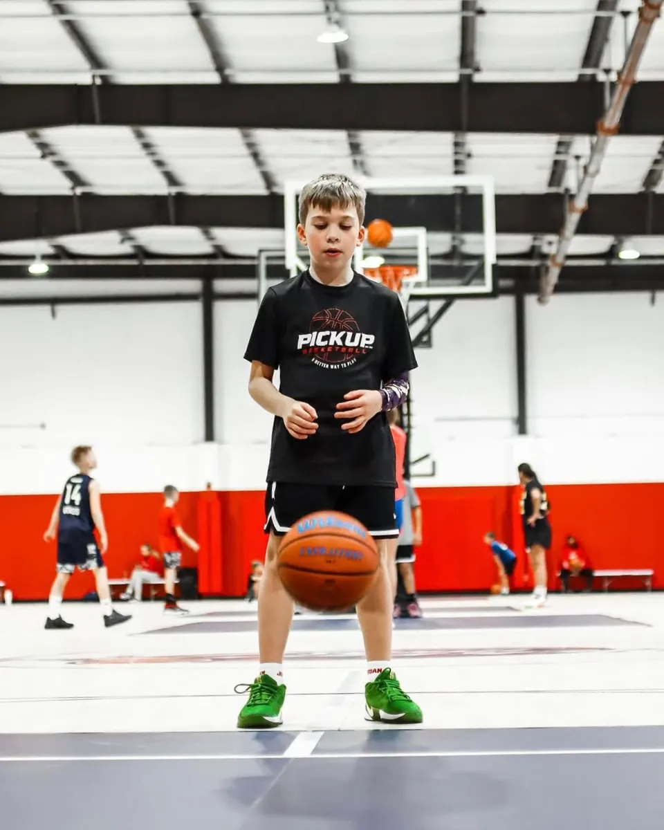 Young boy in green shoes and black 'Pickup Basketball' shirt bouncing a basketball on an indoor court with other kids in the background.