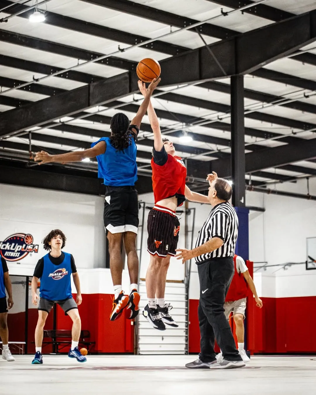 Two basketball players jumping to tip off the ball as a referee tosses it on an indoor court.