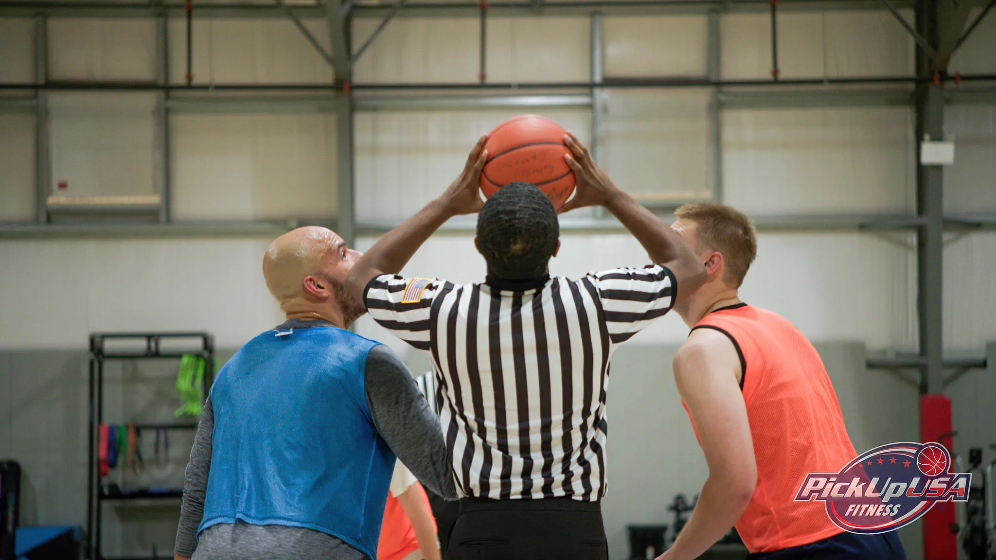 Basketball referee holding the ball above his head preparing for a jump ball between two players in blue and orange jerseys inside a gym.