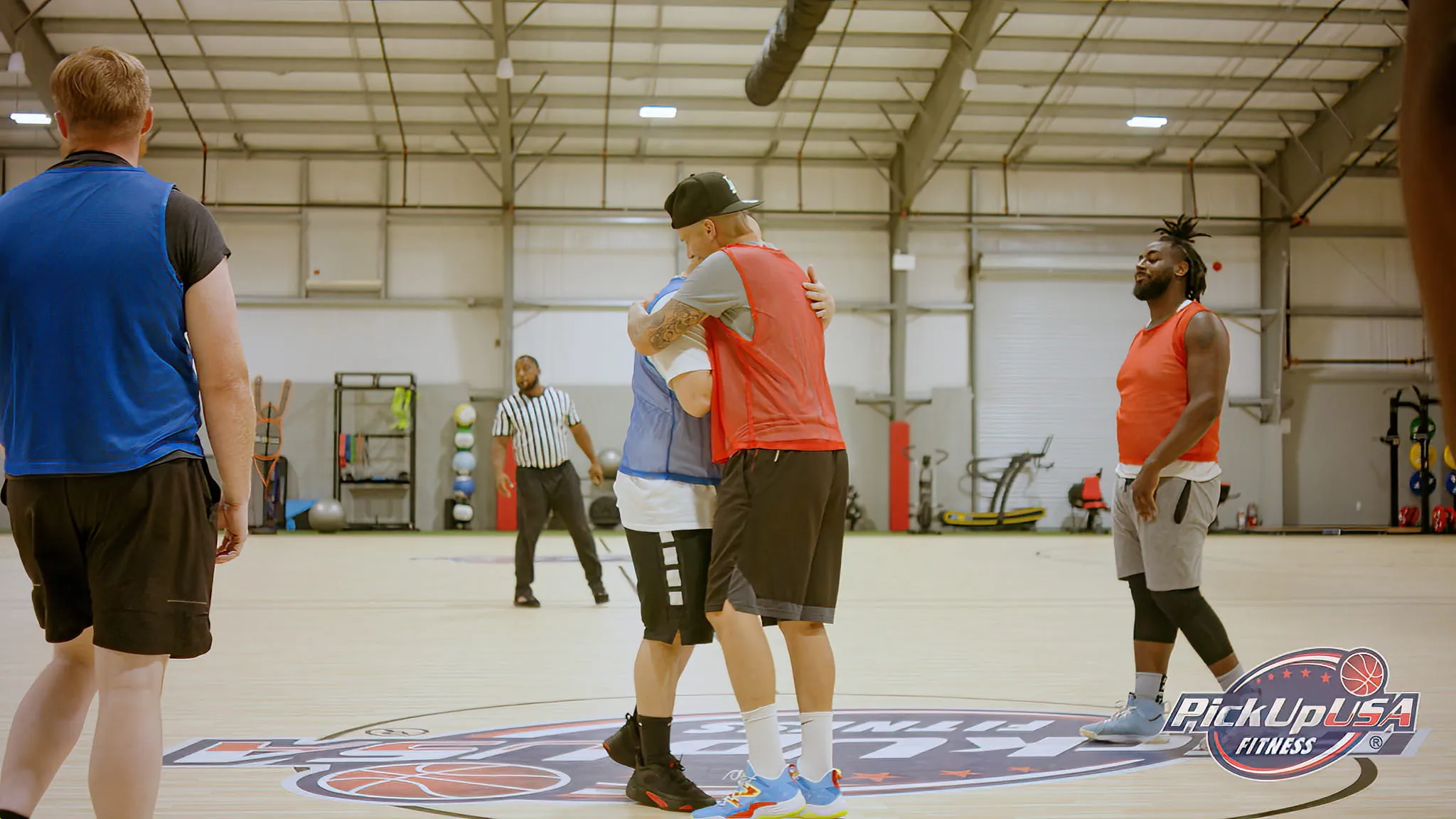 Two basketball players, one in a red jersey and one in a blue jersey, hugging at center court in an indoor gym while others look on.
