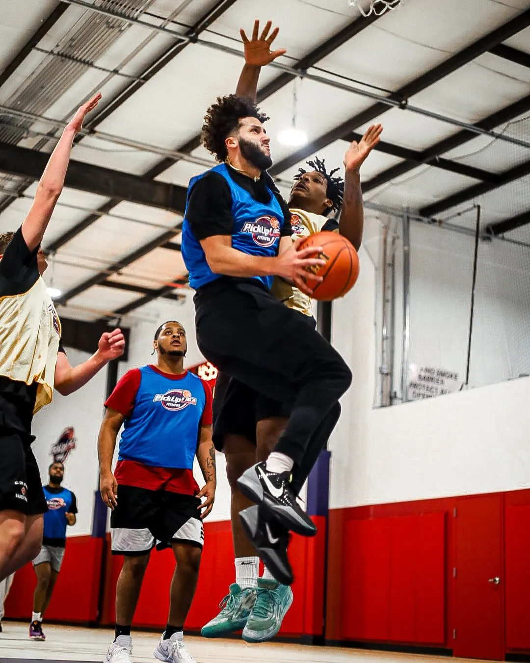 Basketball player in blue jersey jumping for a layup while defended by a player in yellow jersey at indoor court.