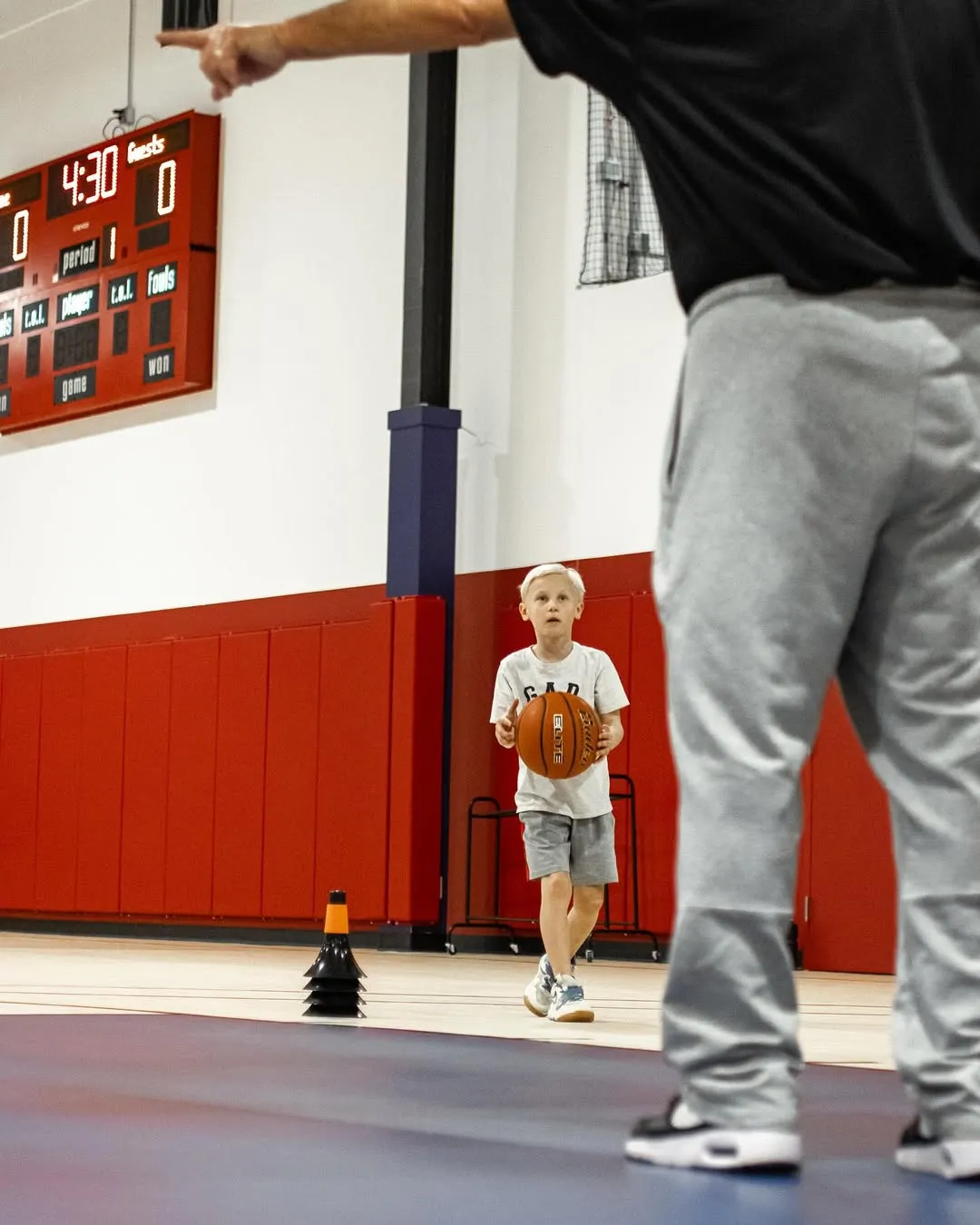 Young boy holding a basketball in a gym with a coach pointing and a scoreboard showing 4:30 left.
