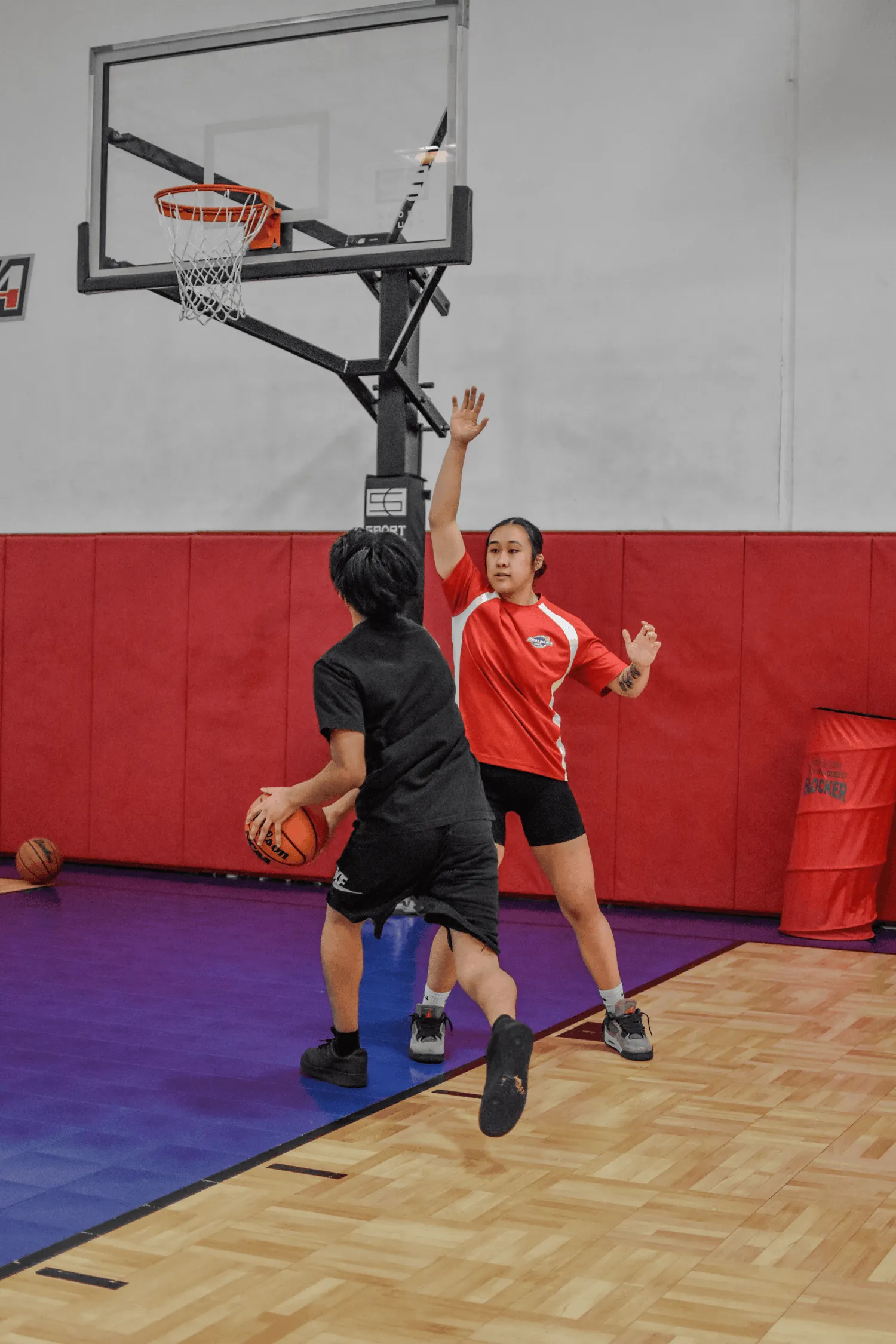 Two young basketball players; one in black holds ball, guarded by player in red jersey on court.