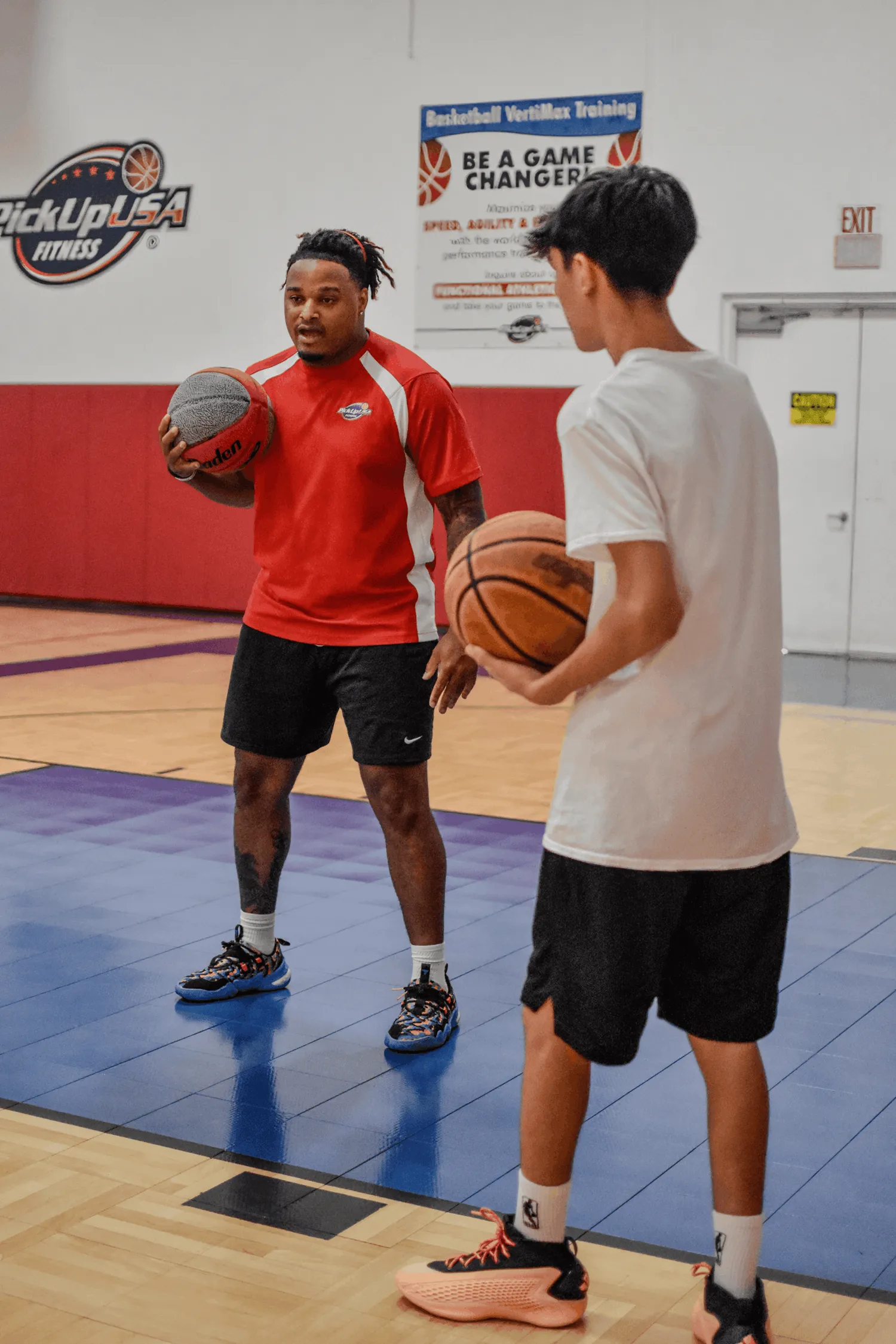 Two men on a basketball court holding basketballs; one wears red shirt, the other white.