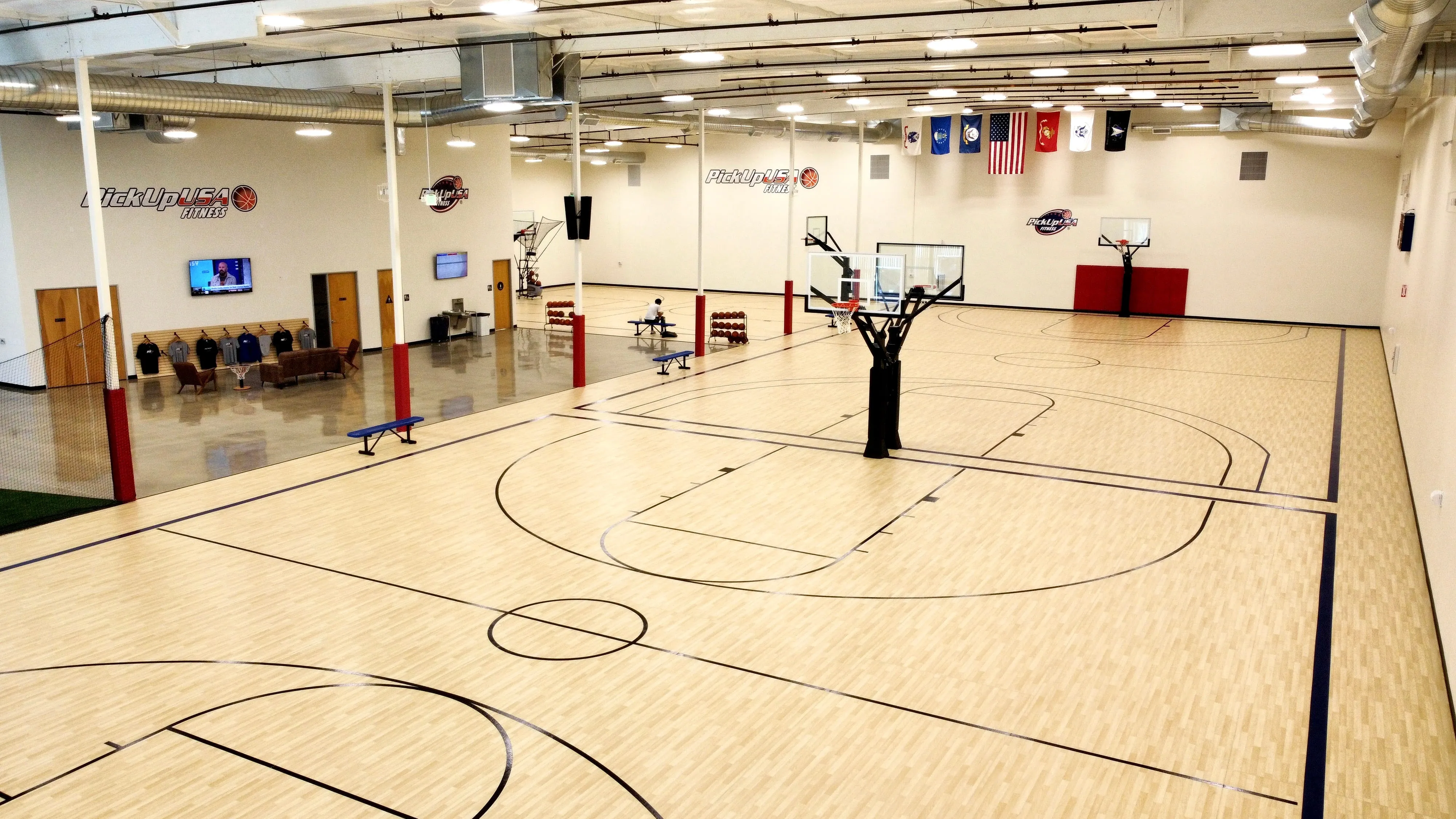 Indoor basketball court with light wood floor, hoops, flags, seating area, and fitness branding on walls.