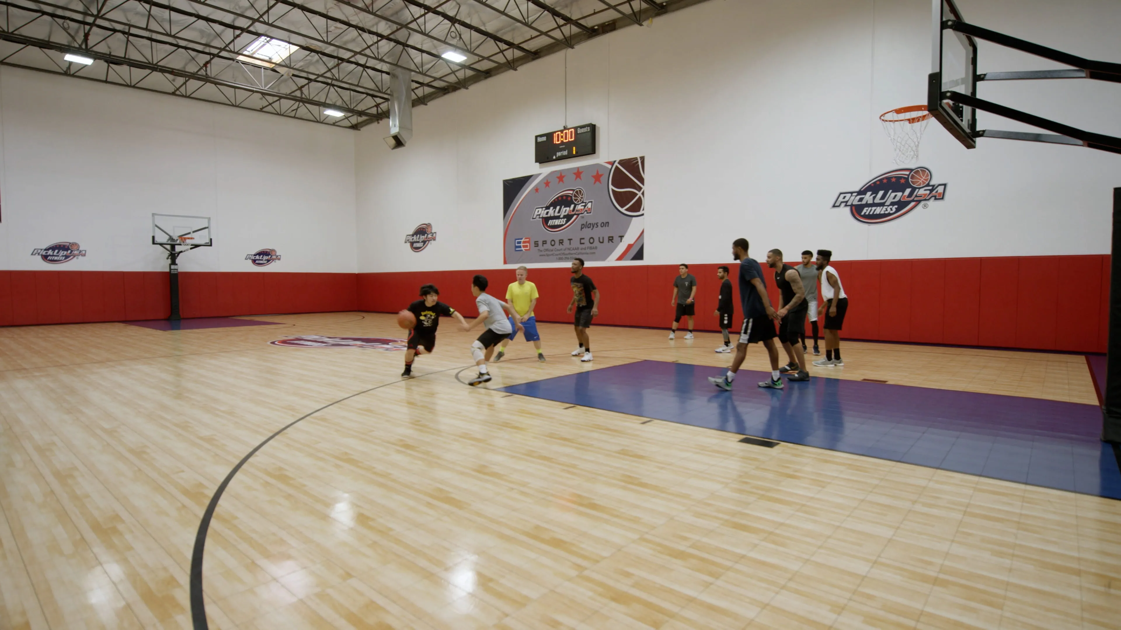 Group playing basketball on an indoor court with PickUpUSA Fitness logos and a scoreboard.
