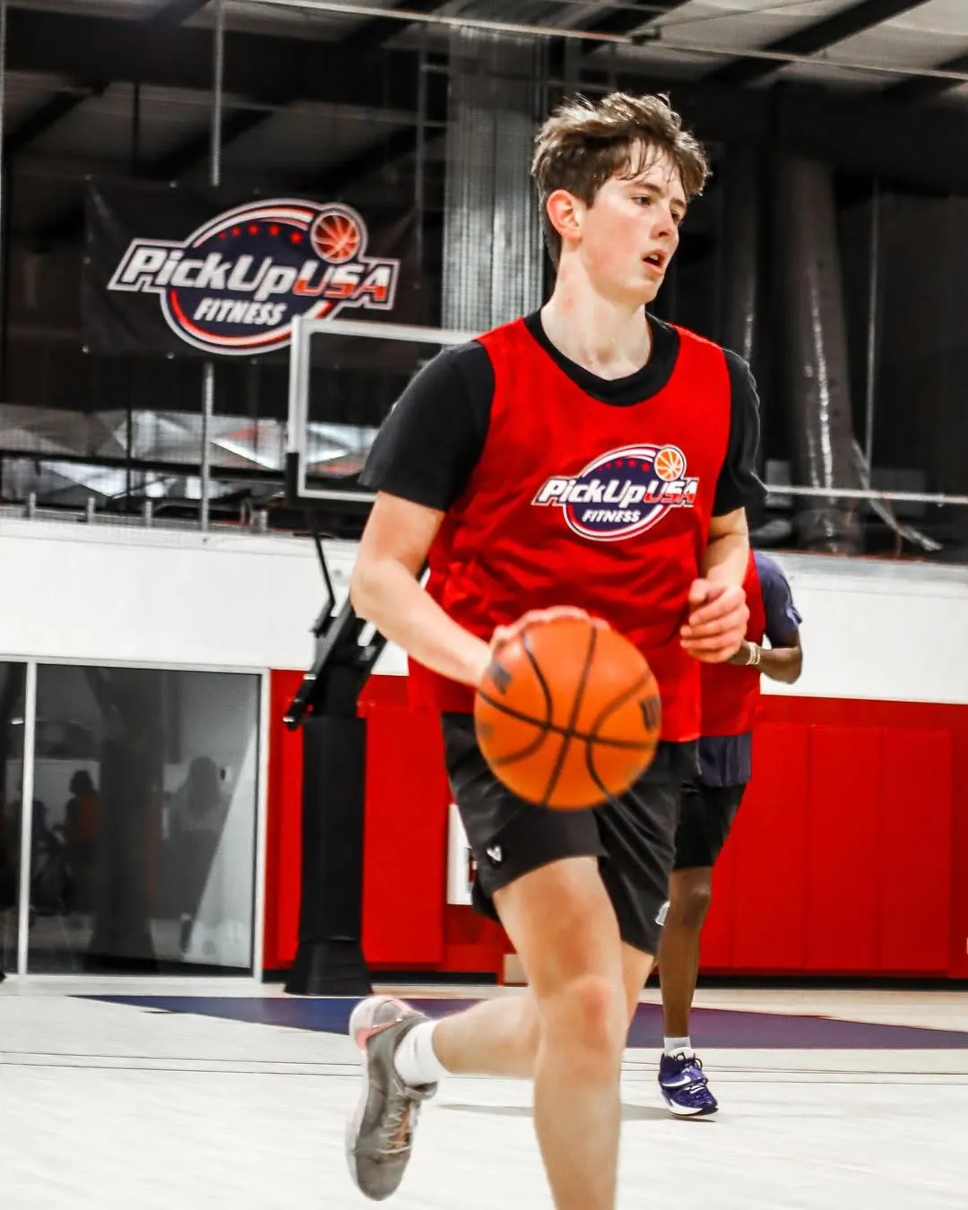 Young male basketball player dribbling on indoor court wearing red PickUp USA Fitness jersey.