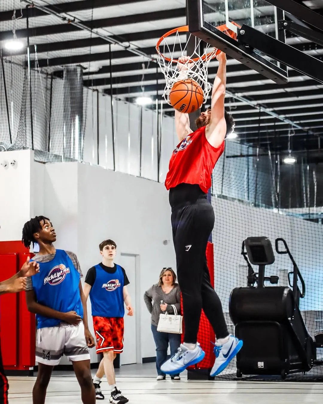 Basketball player in red jumps to dunk while others watch on indoor court.