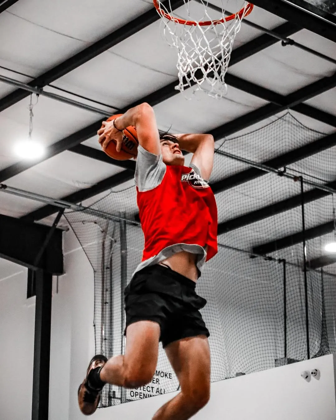 Male basketball player in red jersey jumps to dunk a basketball indoors under the hoop.