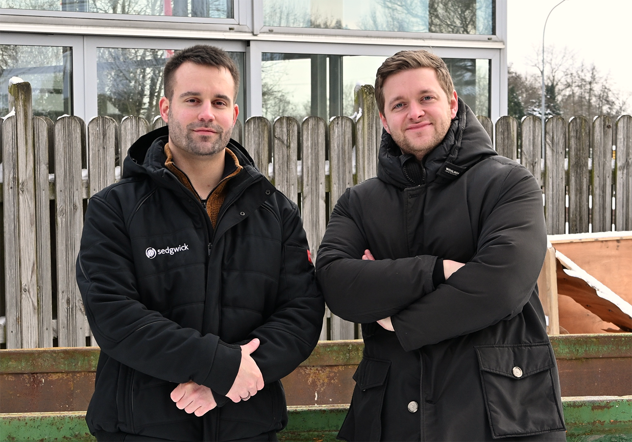 Two men wearing black winter jackets standing outdoors in front of a wooden fence and glass windows.