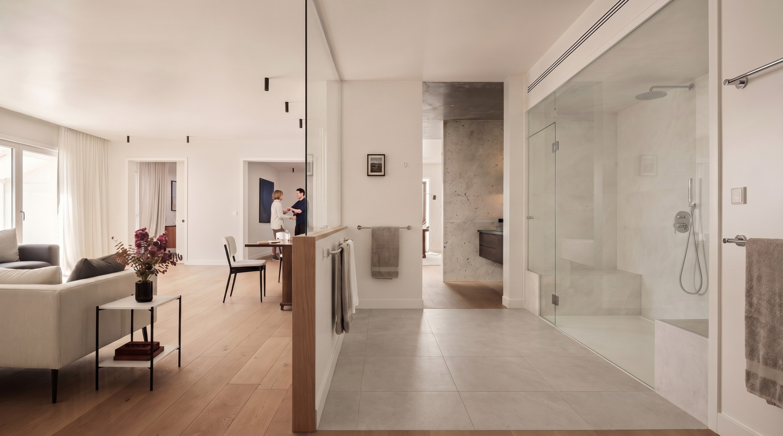 Modern open apartment with a glass shower, wooden floors, gray sofas, and two people talking near the dining table.