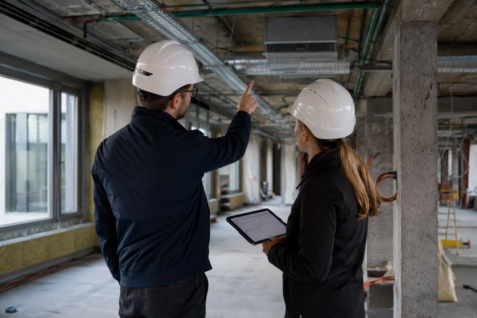 Two construction workers wearing white helmets inspecting an unfinished building interior.