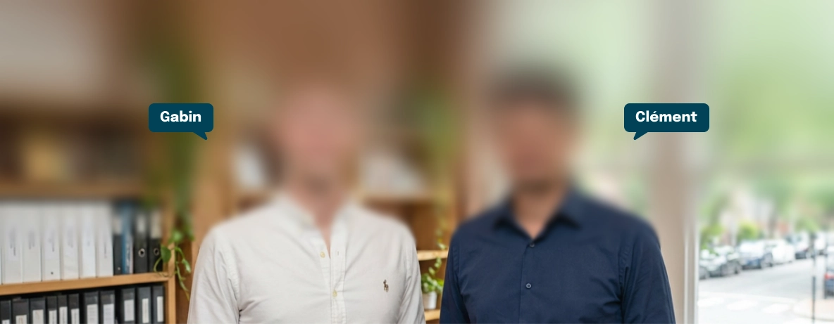 Two men standing indoors in front of a bookshelf filled with binders; the man on the left wears a white shirt and the man on the right wears a dark blue shirt.