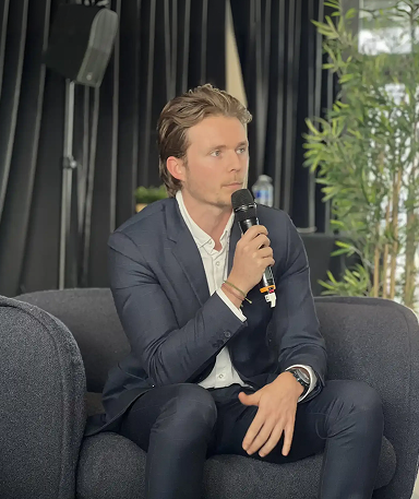 Man in a navy suit sitting on a dark gray chair holding a microphone, speaking or preparing to speak.