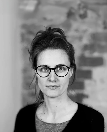 Black-and-white portrait of a woman with glasses and hair tied back, wearing a dark cardigan over a light top, standing in front of a blurred brick wall.