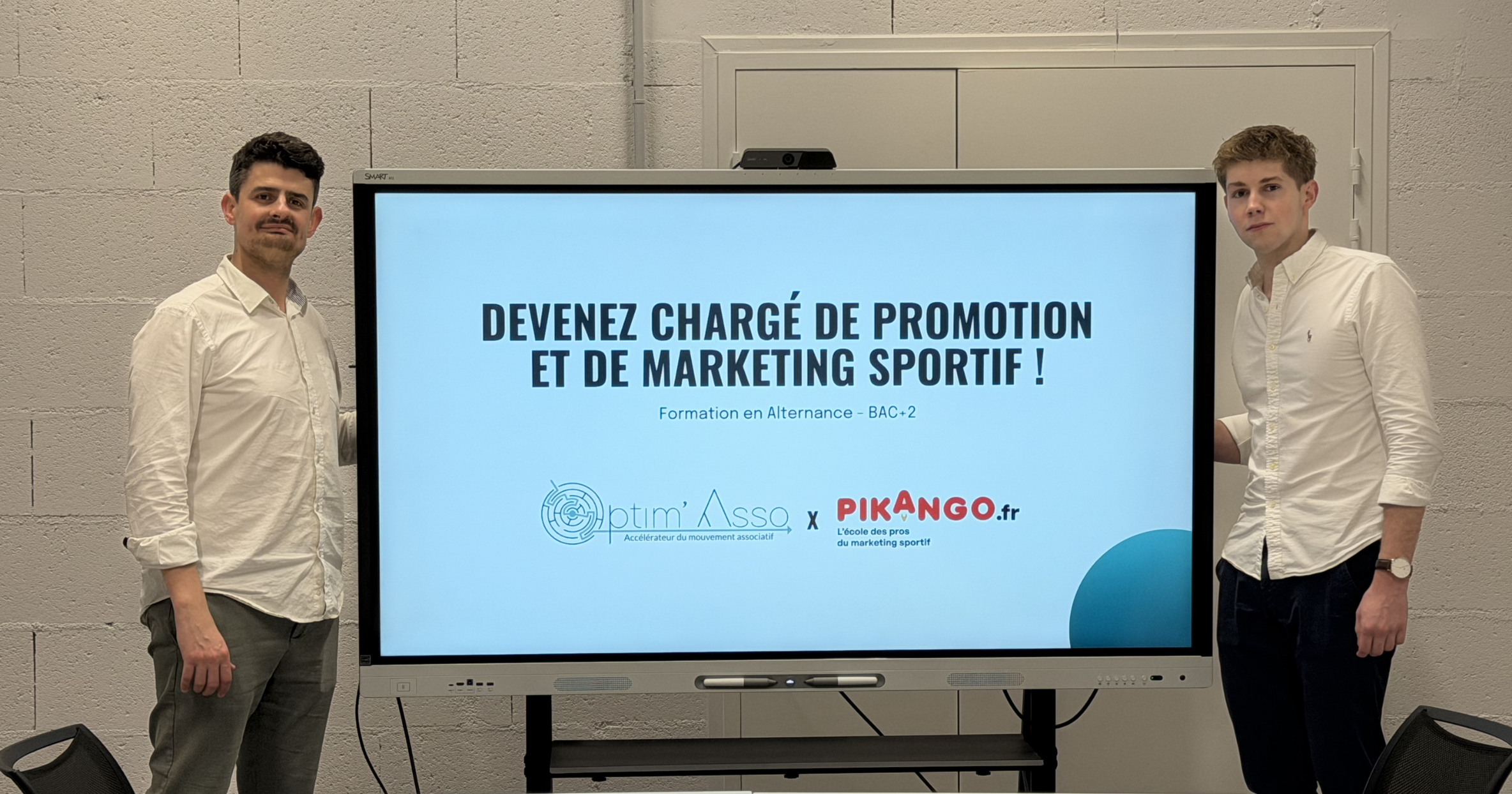 Two men standing indoors in front of shelves and a window, one in a white shirt and the other in a dark blue shirt.