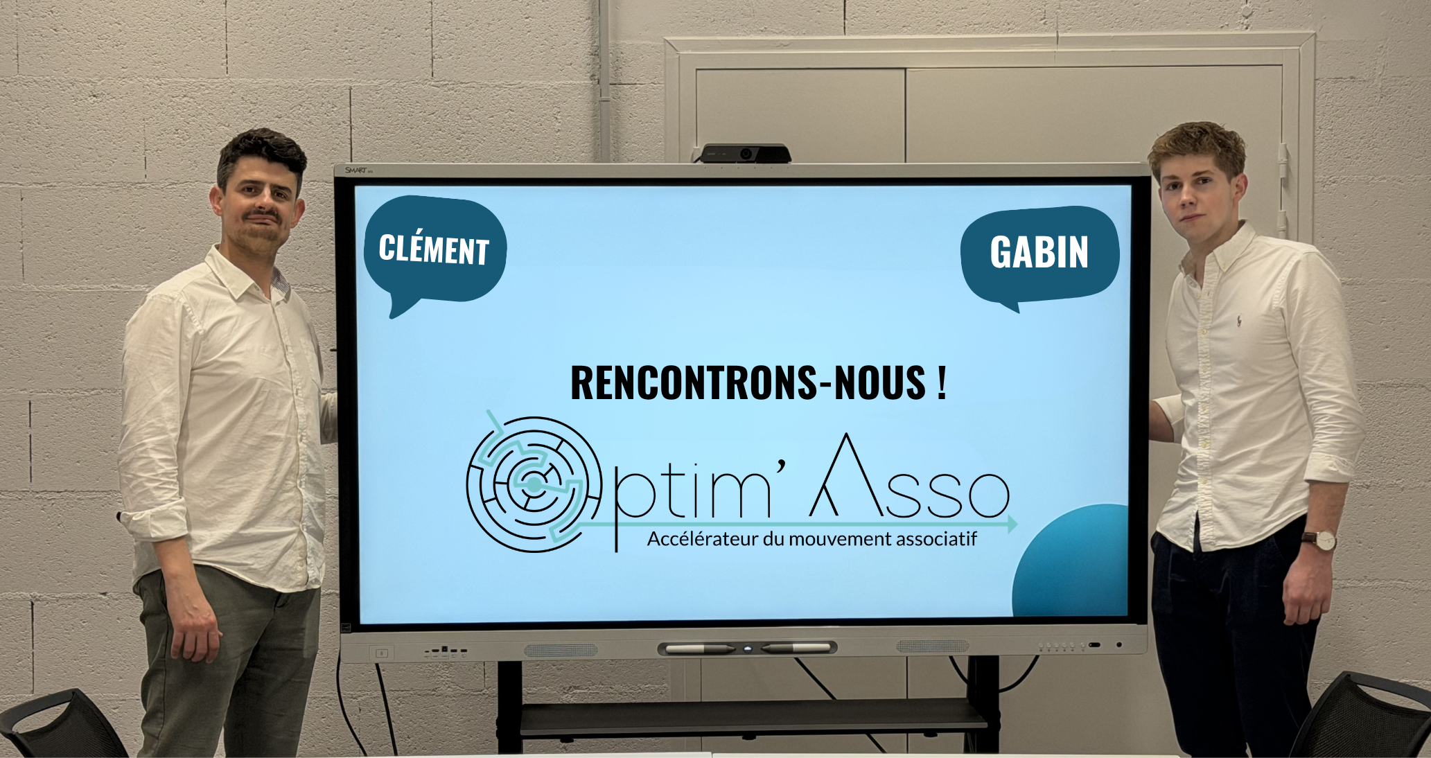 Two men standing indoors in front of a bookshelf filled with binders; the man on the left wears a white shirt and the man on the right wears a dark blue shirt.