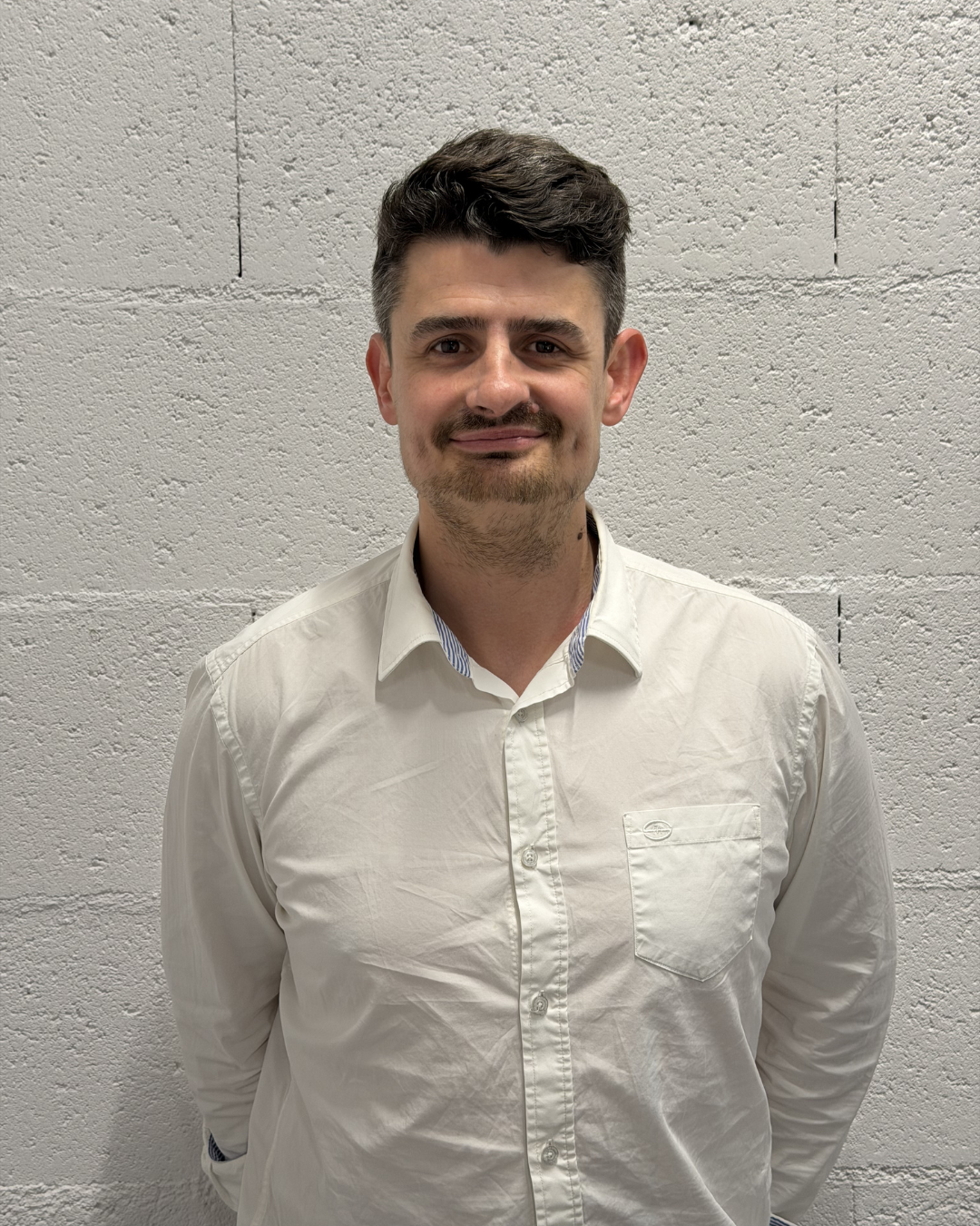 Young man with short dark hair and a trimmed beard wearing a white collared shirt, looking at the camera against a white background.