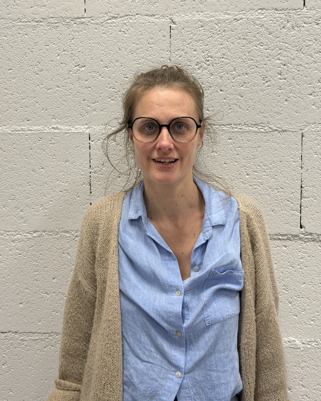 Black-and-white portrait of a woman with glasses and hair tied back, wearing a dark cardigan over a light top, standing in front of a blurred brick wall.