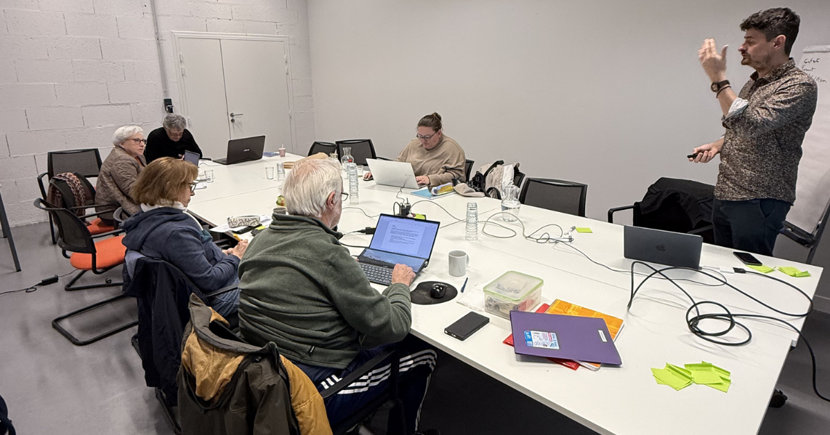 Business meeting with six people around a conference table with laptops and gift bags, while one person stands presenting.