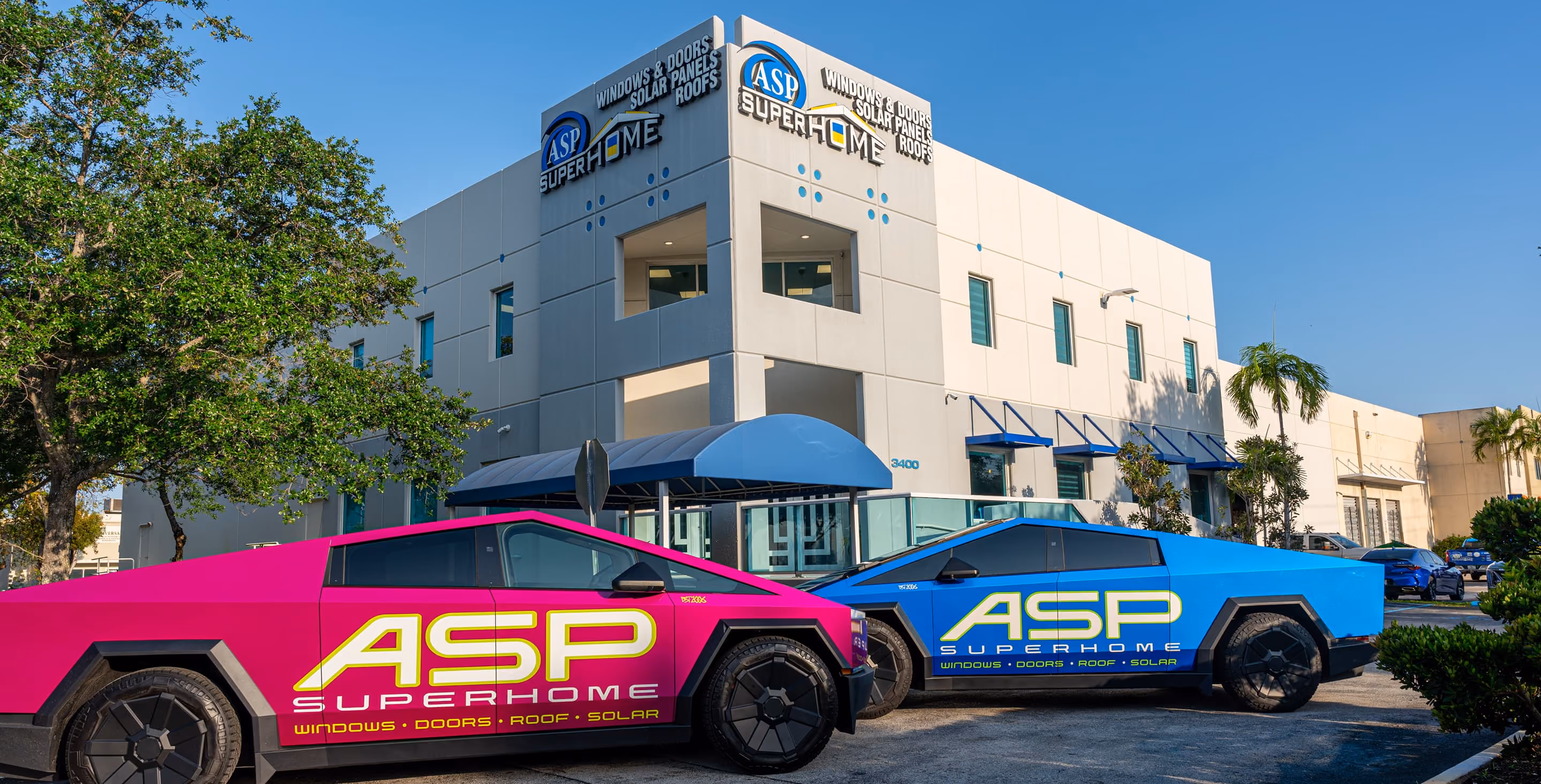 Two futuristic electric pickup trucks, one pink and one blue, with ASP SuperHome branding parked in front of a commercial building with ASP SuperHome signs.