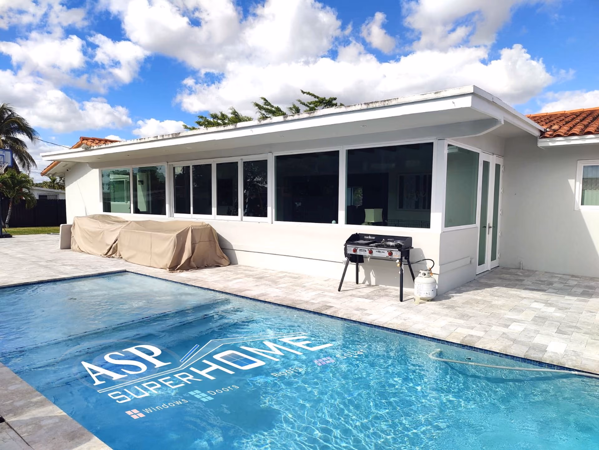 Backyard view of a modern house with large windows, a covered outdoor grill, and a clear swimming pool reflecting the sky.