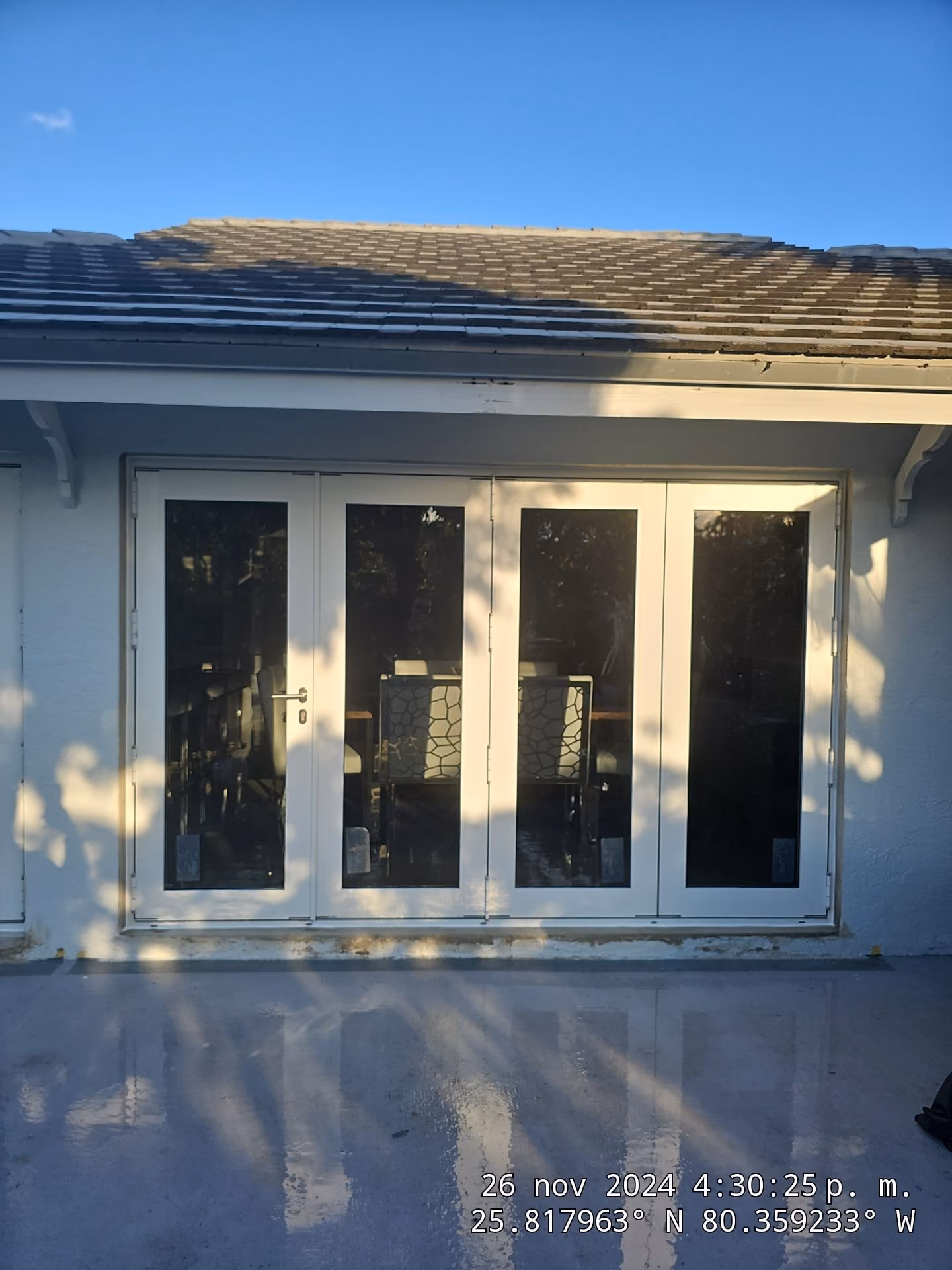 White-framed residential impact doors reflecting shadows and a clear sky, with chairs visible inside.