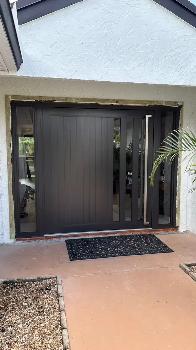 Modern black sliding front door with narrow vertical glass panels on each side and a large metallic handle, set in a light textured wall with a black decorative doormat.
