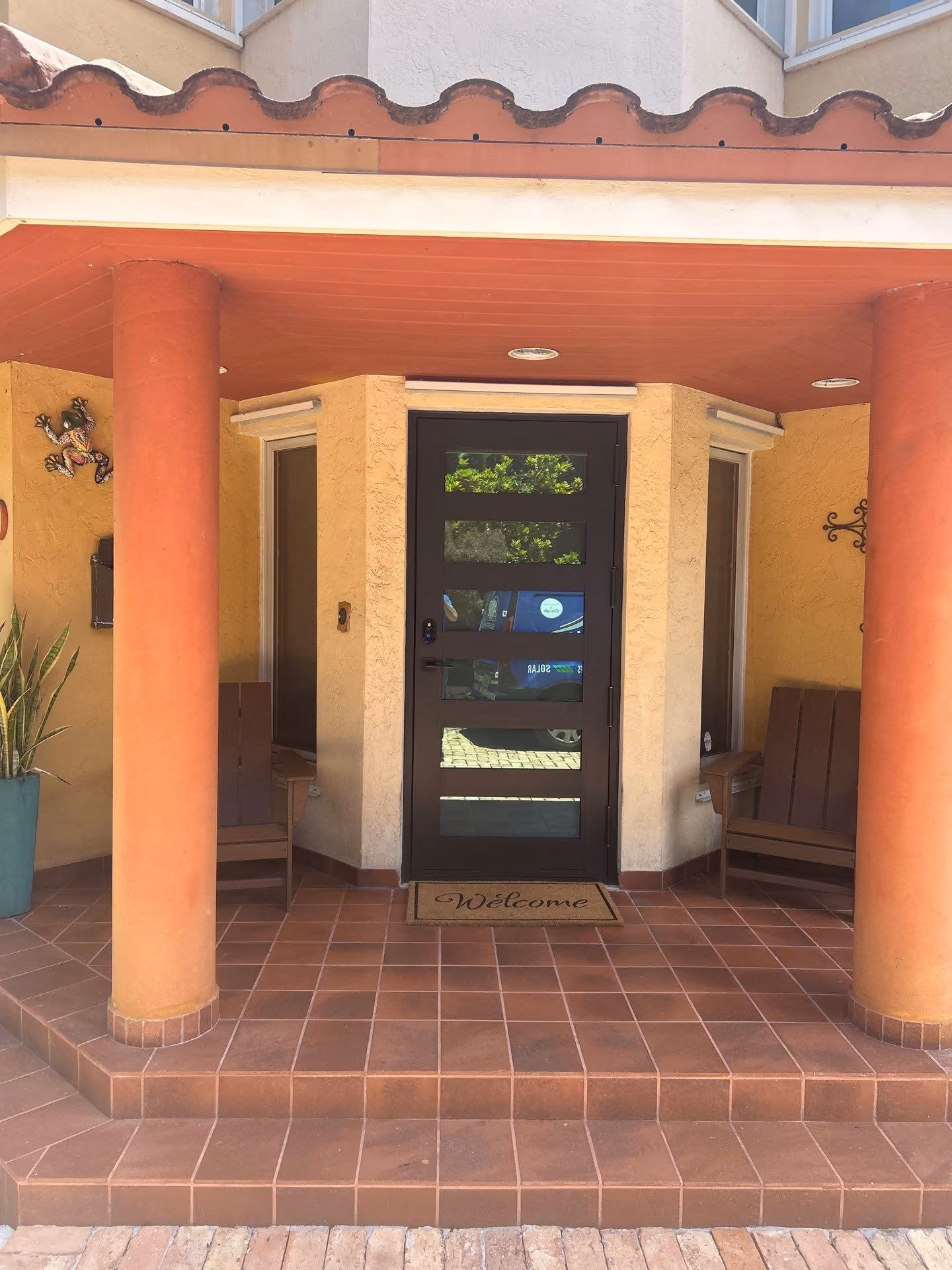 Front entrance with a black door featuring horizontal glass panels, flanked by two orange columns and two brown chairs, with a terracotta tiled floor and a welcome mat.