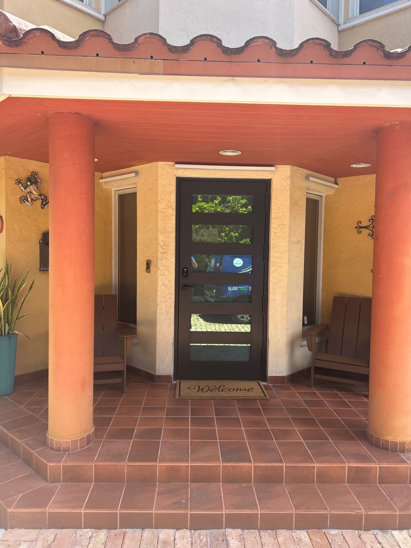 Front entrance with a black door featuring horizontal glass panels, flanked by two orange columns and two brown chairs, with a terracotta tiled floor and a welcome mat.