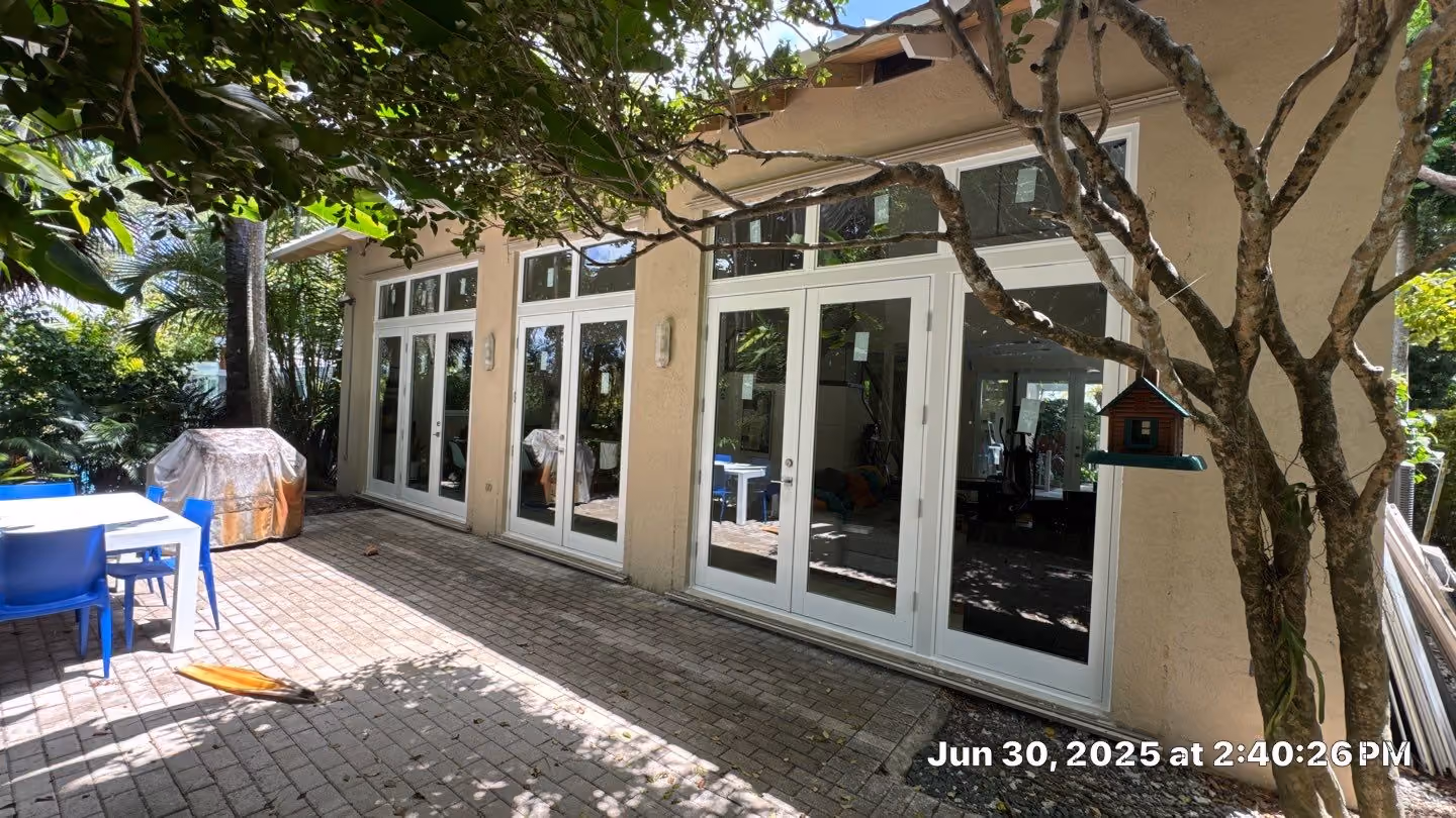 Patio area with white double glass doors, blue chairs around a white table, a covered grill, and tree branches casting shadows.