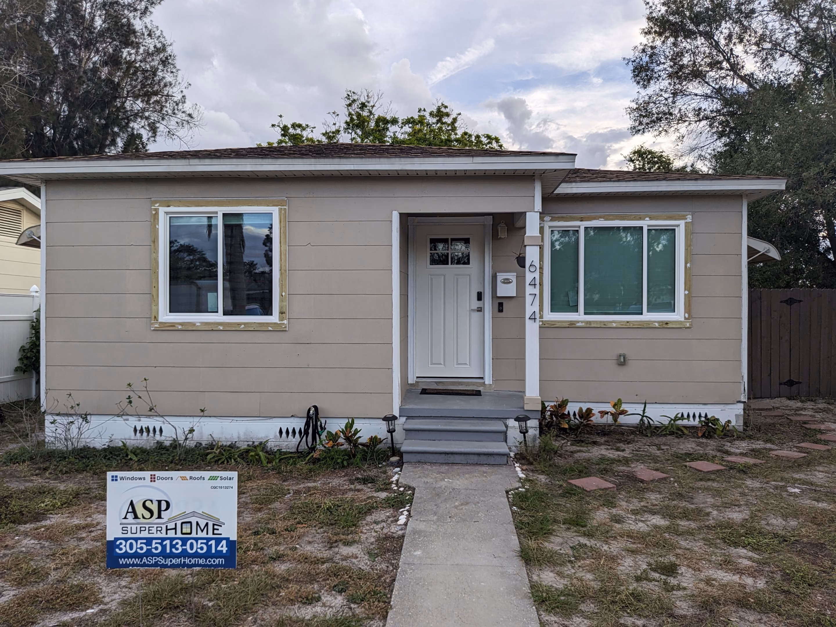 Single-story beige house with white front door and two windows, a sidewalk leading to the entrance, and a yard sign advertising ASP Super Home services.
