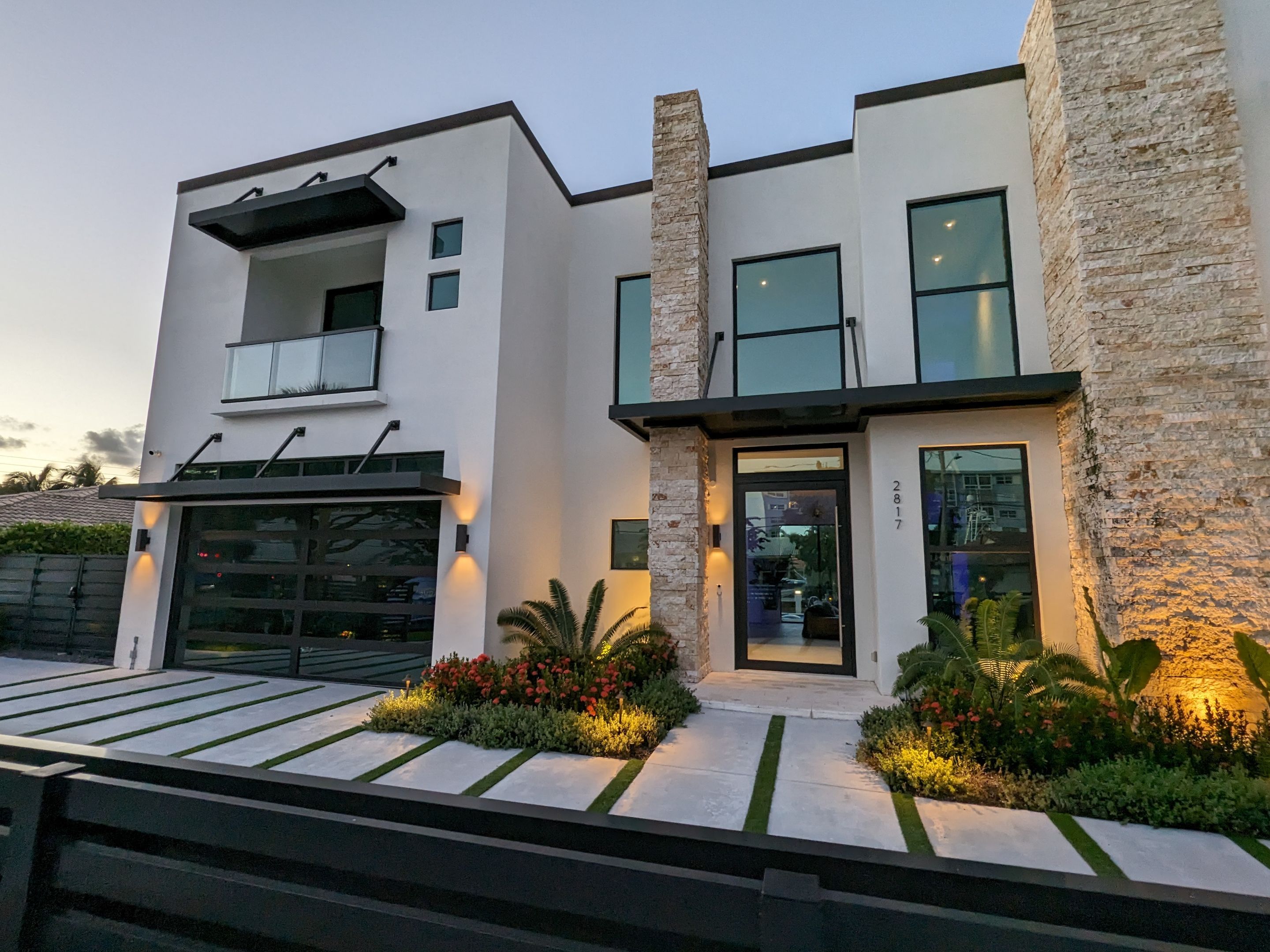 Modern two-story house with white exterior, stone accents, large windows, illuminated landscaping, and a driveway with grass strips.