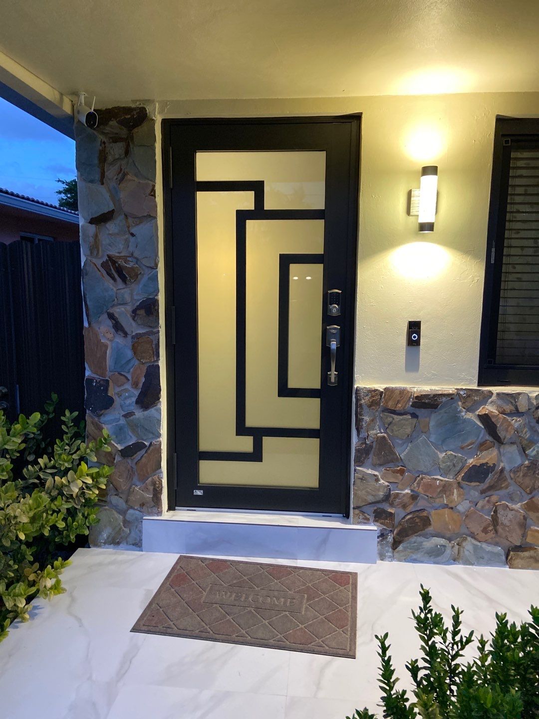 Modern french door with frosted glass and geometric black frame, stone walls, welcome mat, and illuminated wall light.
