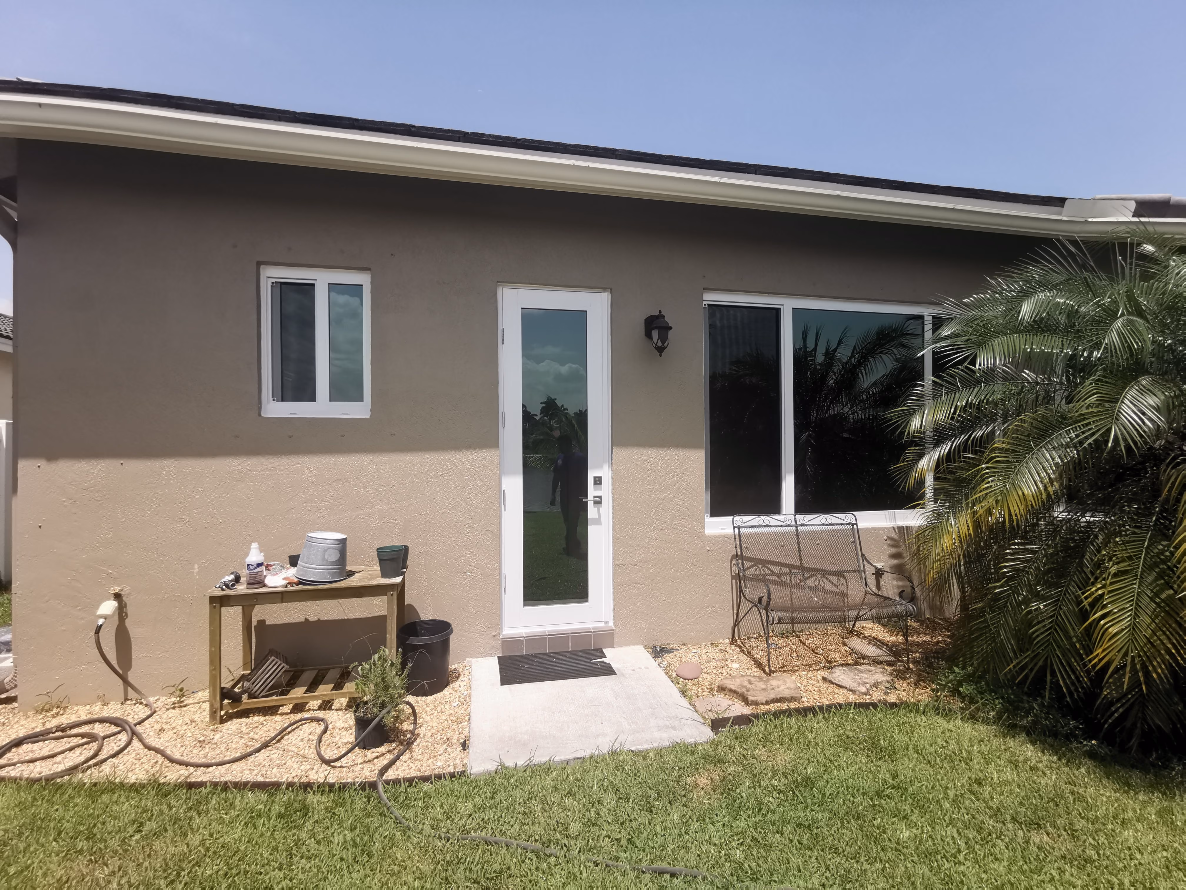 Backyard view of a beige house with a white glass door, small window, metal bench, potted plants, and a garden hose on green grass.