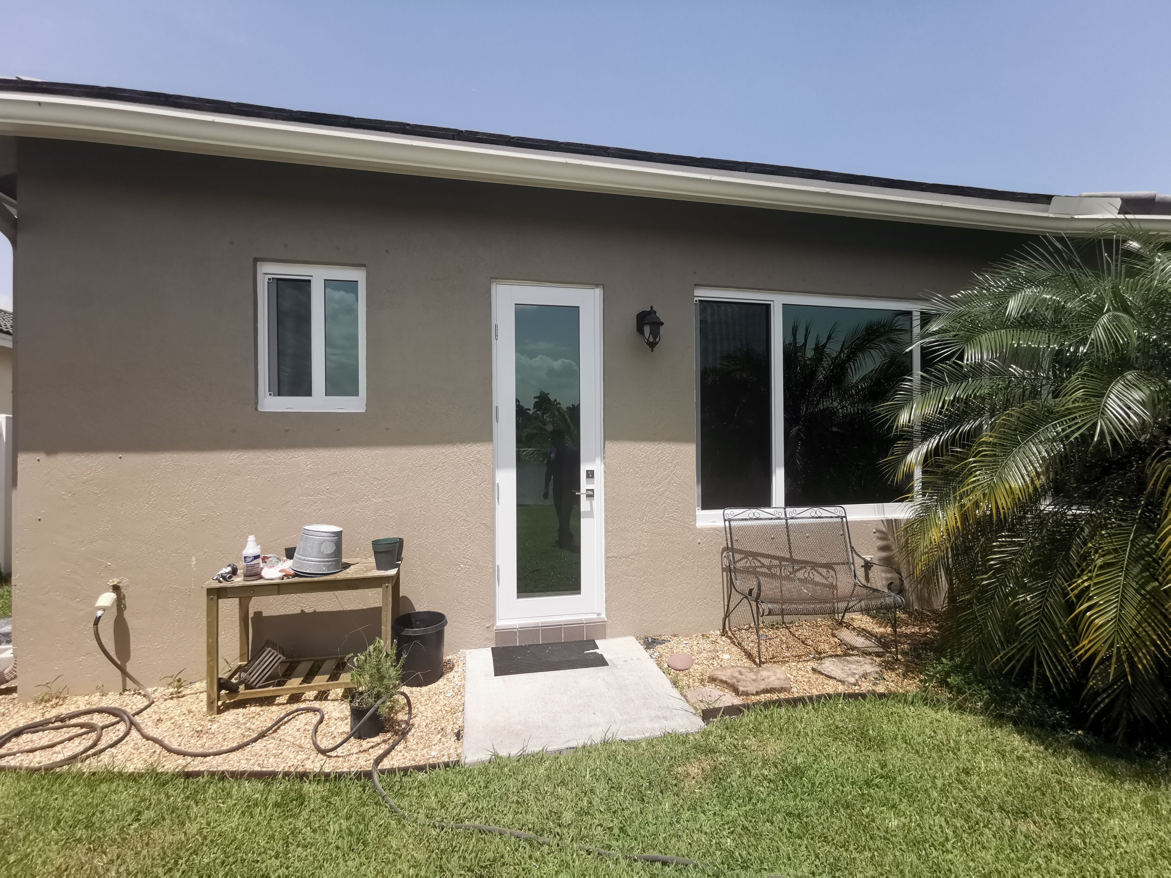 Backyard view of a beige house with a white glass door, small window, metal bench, potted plants, and a garden hose on green grass.