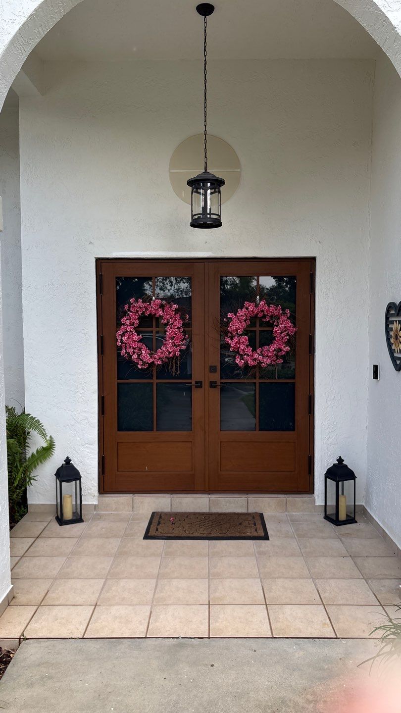 Front porch with double french doors decorated with two pink floral wreaths, flanked by black lanterns on tiled floor, under a hanging black lantern light.