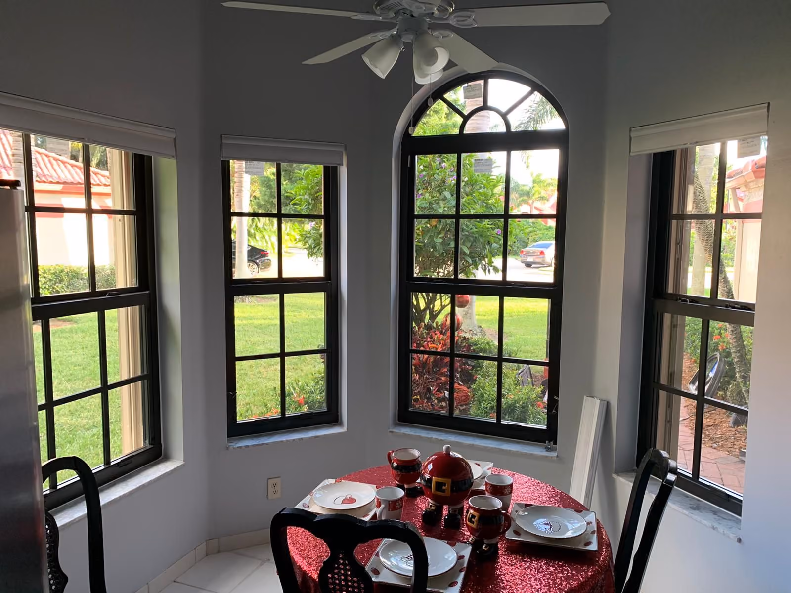 Dining area with a round table covered in a red sequined cloth set for four, surrounded by windows showing green lawn and outdoor plants.