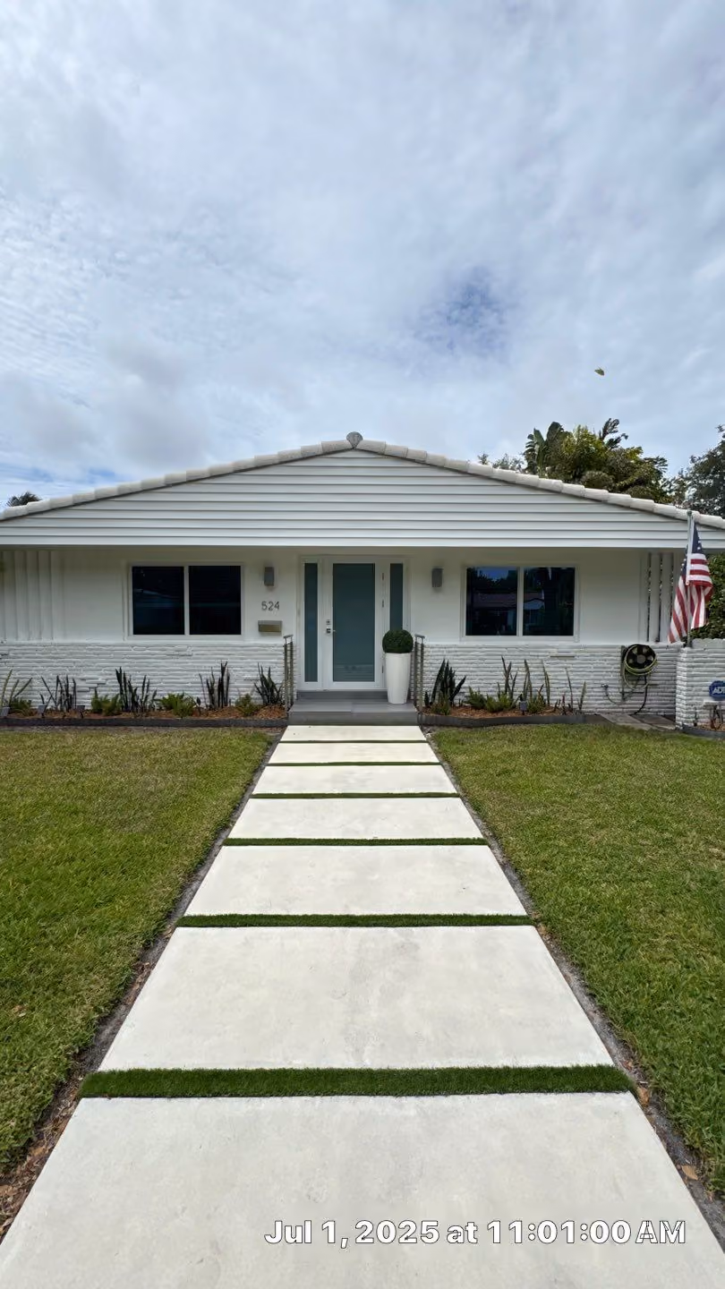 Single-story white house with a modern concrete and grass pathway, green lawn, two front windows, a blue impact door, and an American flag on the right side.