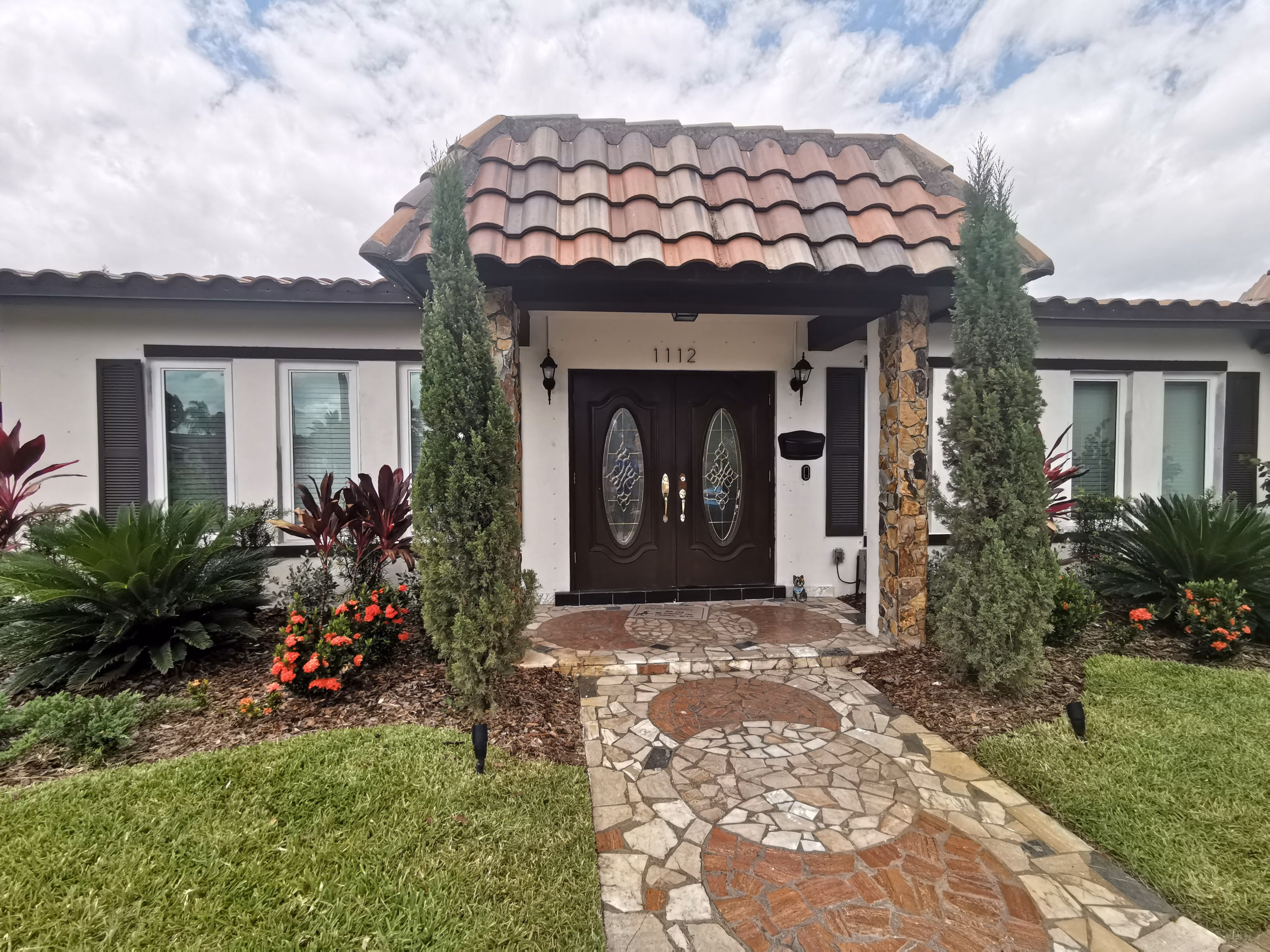 Front entrance of a house with dark wooden double impact doors, stone tile walkway, and landscaped garden with tall bushes and colorful flowers.