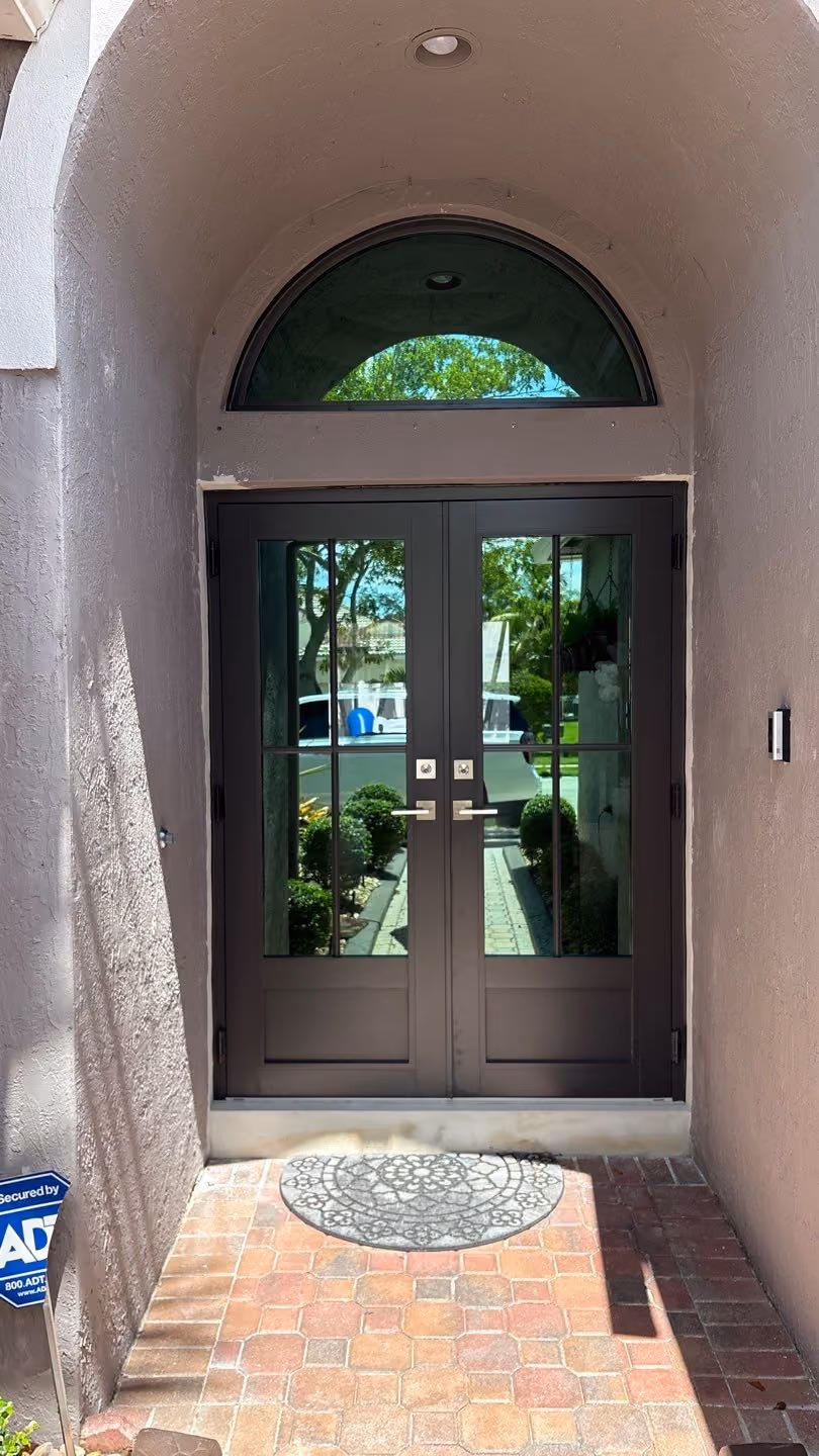 Double black glass impact doors under a curved archway with a decorative gray doormat on a brick pathway.