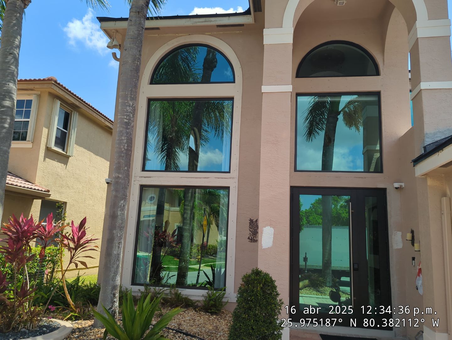 Front entrance of a beige stucco house with large arched and rectangular residential impact windows reflecting palm trees and blue sky, surrounded by tropical plants.