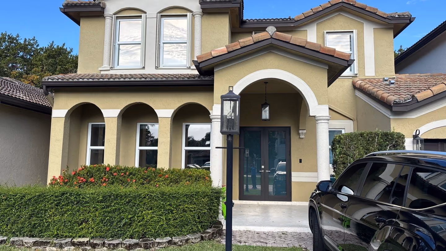 Two-story beige house with arched porch, decorative bushes, and a black SUV parked in front.