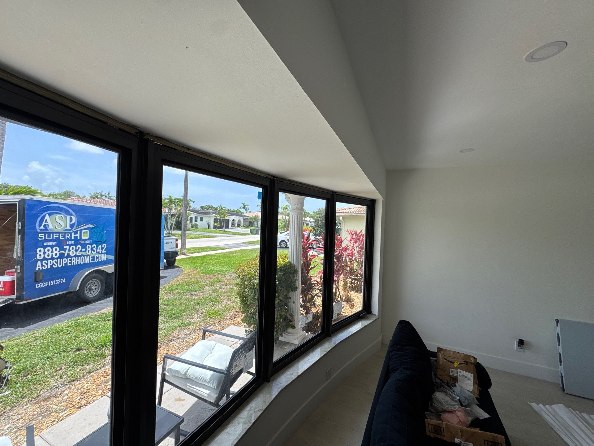Interior view of a room with large black-framed sliding windows looking out onto a green lawn and street, with a blue ASP Superhome trailer parked outside.