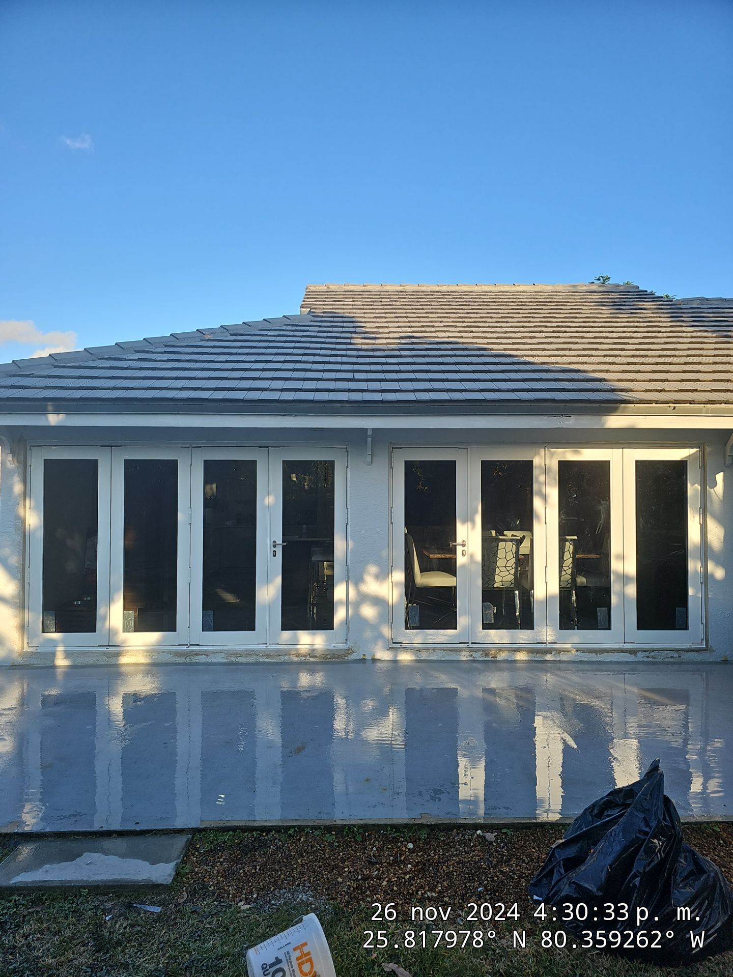 Back of a house with white framed glass doors reflecting sunlight on a wet concrete patio under a clear blue sky.